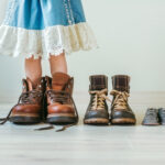 Close up of little girl putting on fathers hiking shoes. Three pairs of shoes for family indoor.